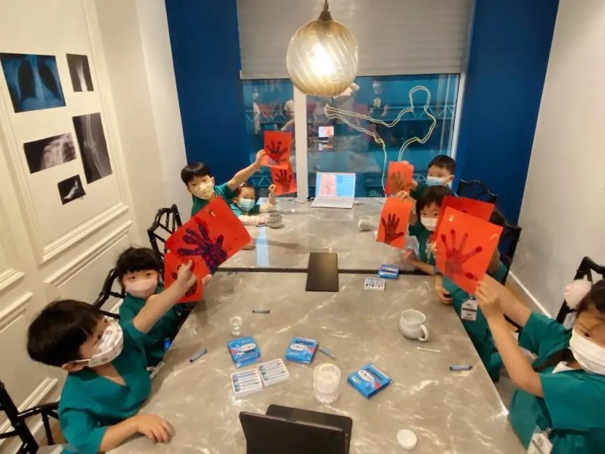 幾位穿著綠色小醫生制服的兒童，圍坐在桌邊舉起紅色手掌形狀的紙板，牆上貼有X光片，體驗X光概念相關的創意活動，是暑假特別優惠的親子主題活動。